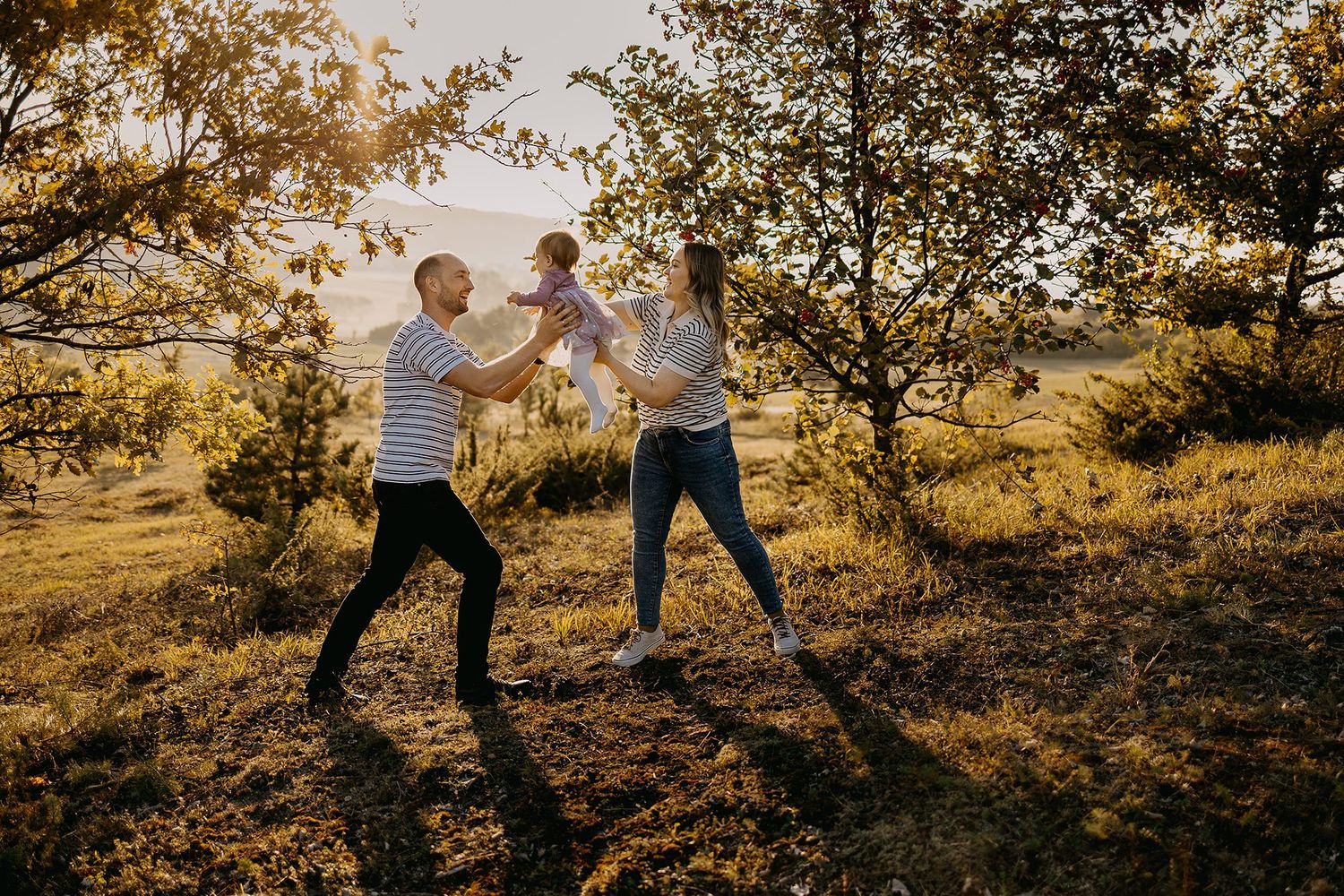 Parents playfully lifting baby together under golden autumn trees at sunset.