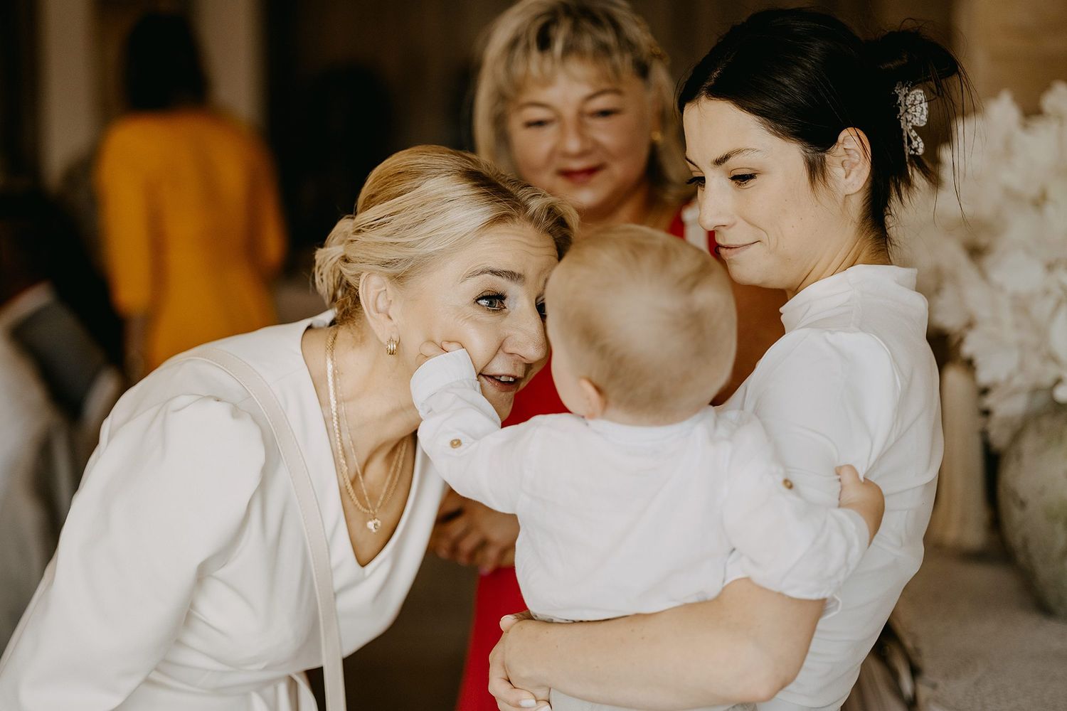 Two women in white outfits lovingly interacting with a baby at a christening celebration outdoors.