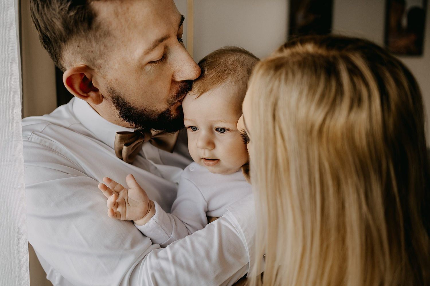 A father kisses his baby while mother looks on during a family christening celebration.