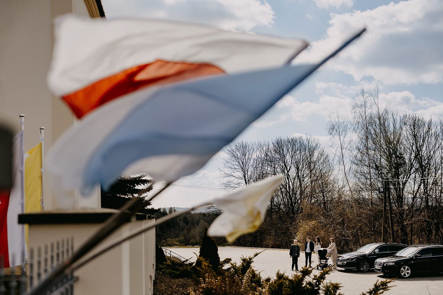Polish flags waving in breeze outside building with people gathered in parking lot on cloudy day.