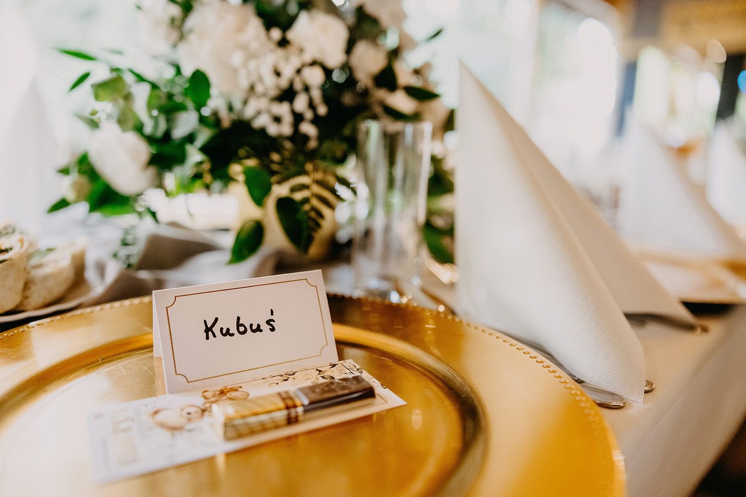 Gold charger plate with Kubus name card and white floral centerpiece at an elegant baptism reception table.