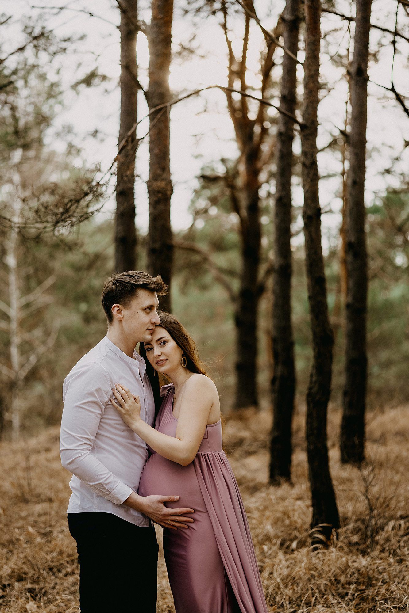Couple embracing in pine forest at sunset, pregnant woman in pink dress cradling her baby bump.