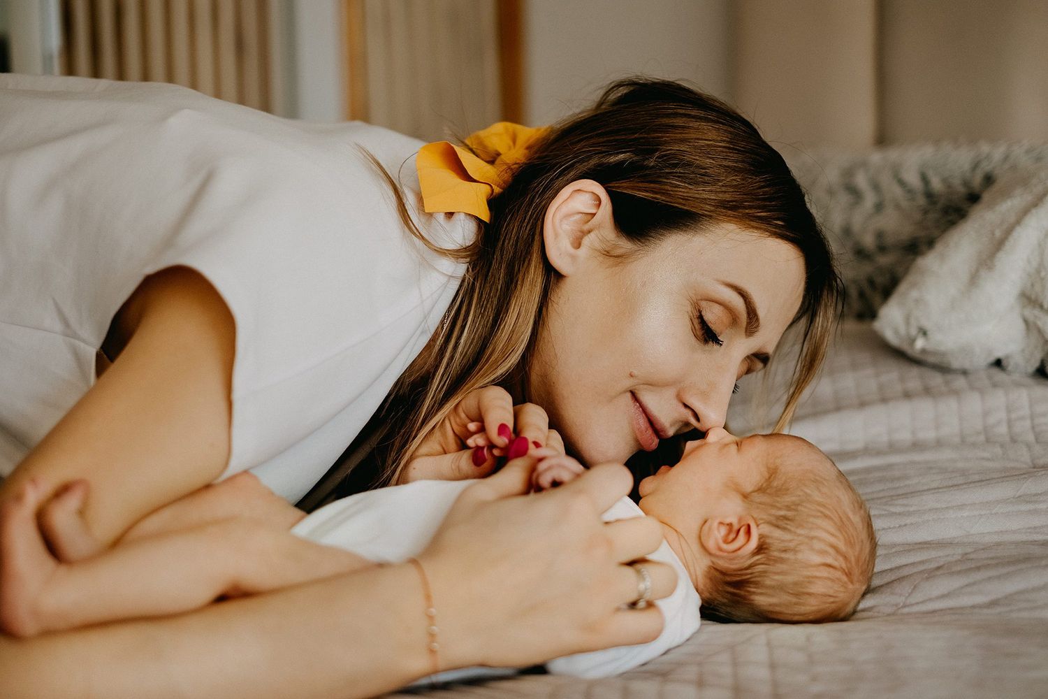 A tender mother lies on a bed, gently nuzzling her sleeping newborn baby.