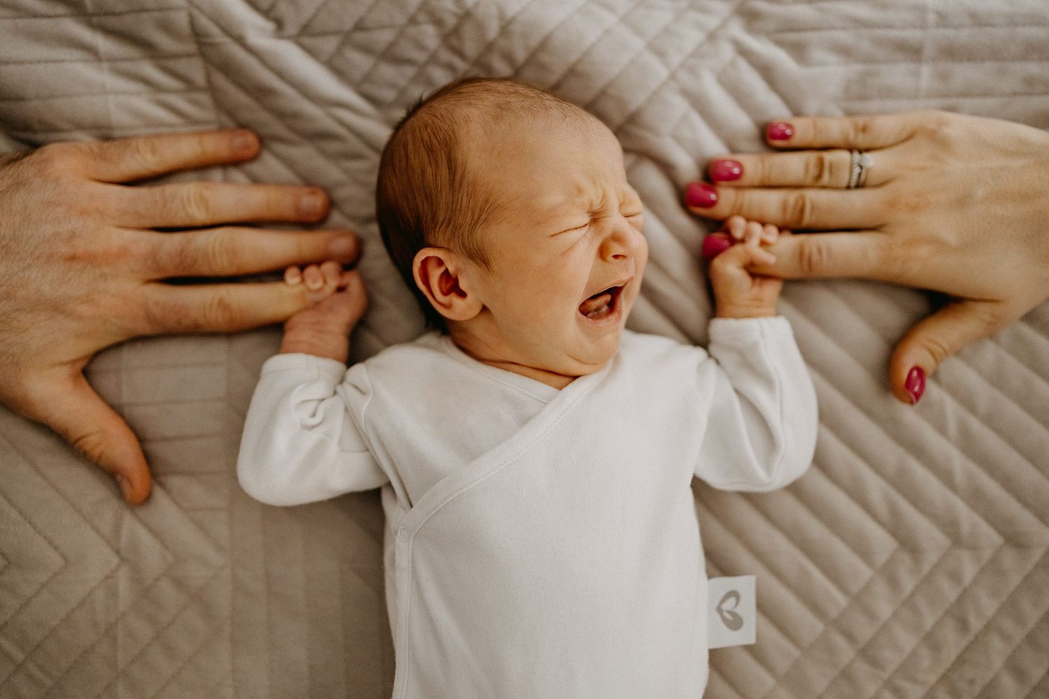 Newborn baby crying while lying on a blanket with parents hands gently holding its arms.