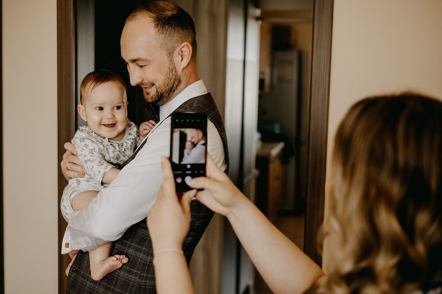 Smiling father holds laughing baby while woman photographs them on smartphone indoors.
