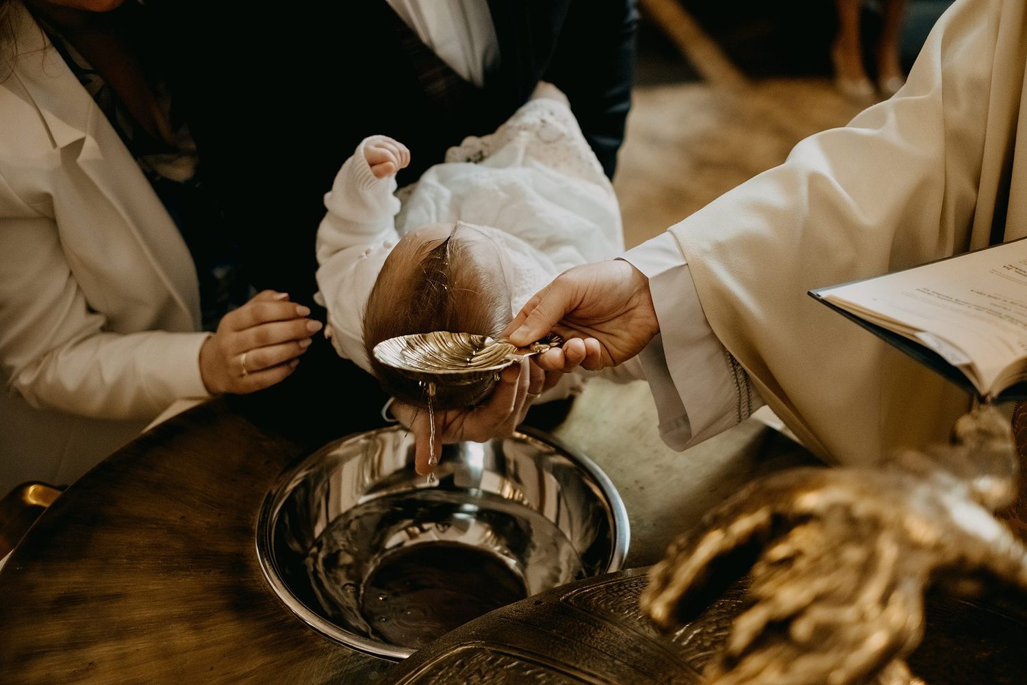 A priest performs a baptism ceremony, pouring holy water over an infant's head at a ornate baptismal font.