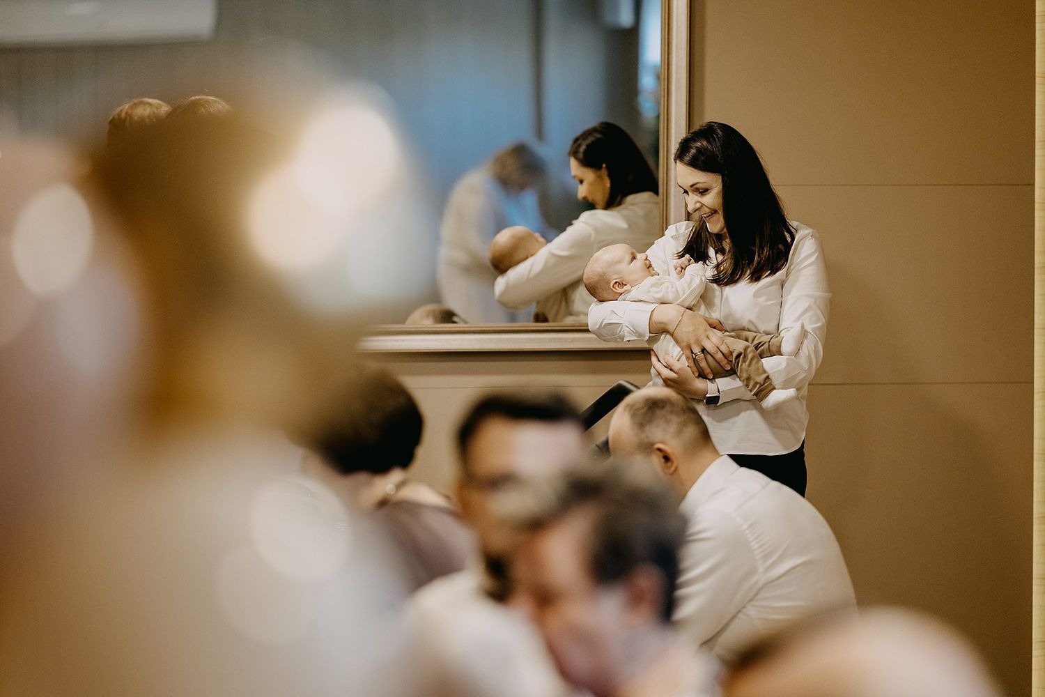 A smiling woman in black holds a baby while seated among guests at a christening event.