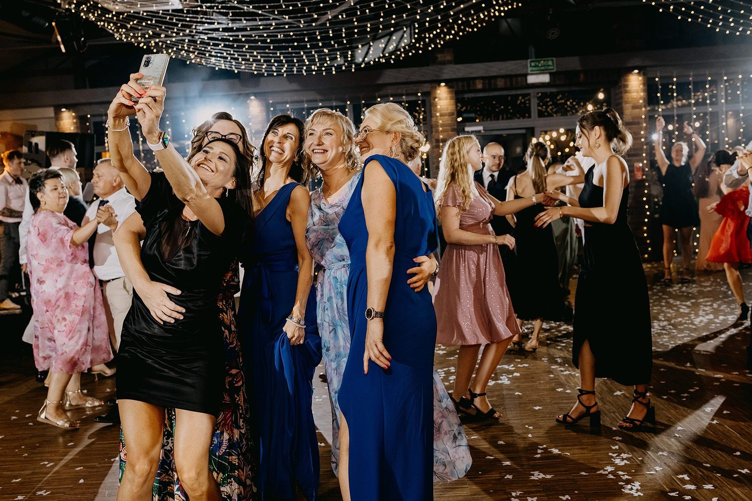 Wedding guests taking group selfie on dance floor surrounded by confetti and string lights.