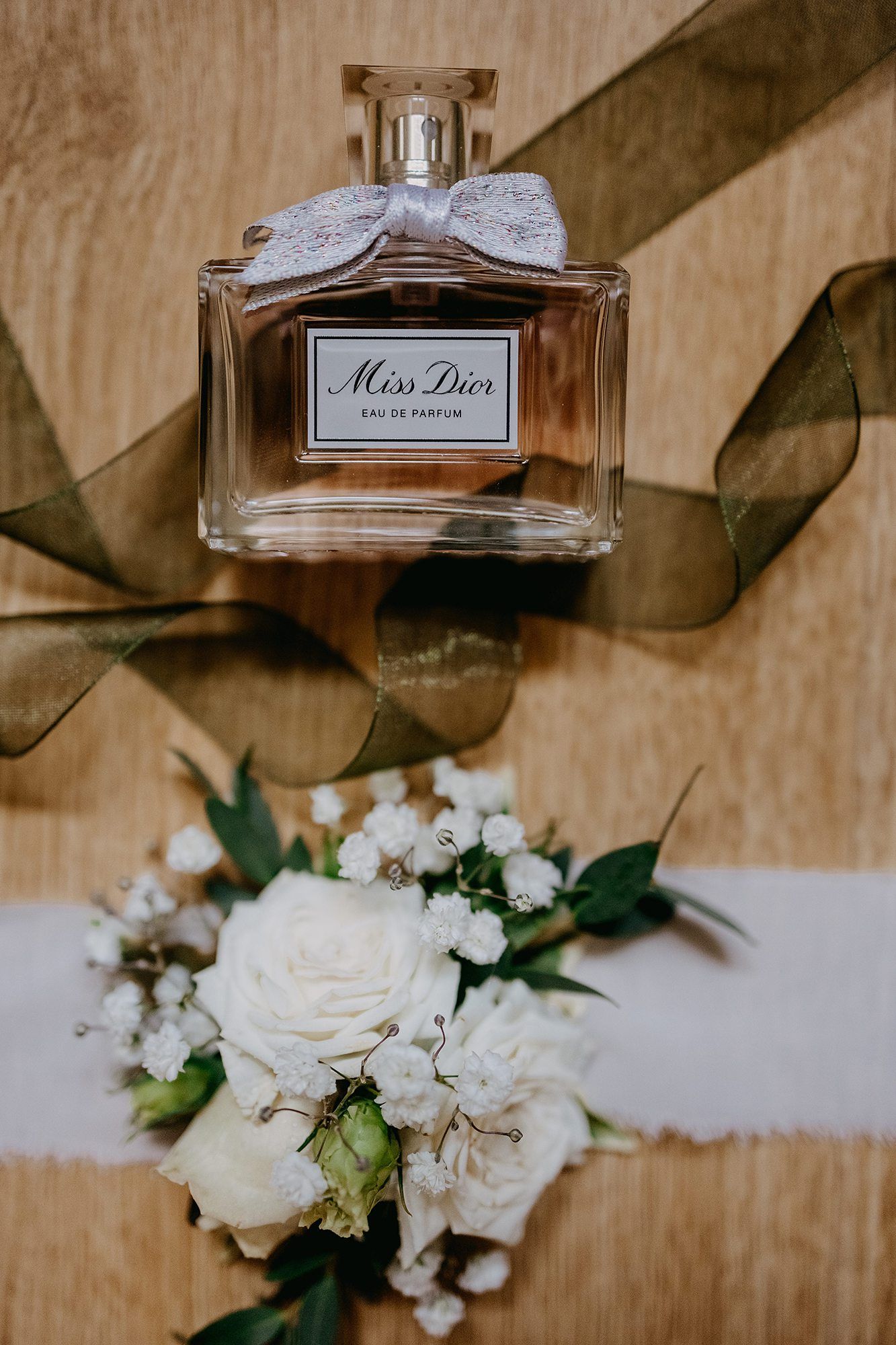 Miss Dior Eau de Parfum bottle with ribbon bow beside white rose boutonniere on wood.