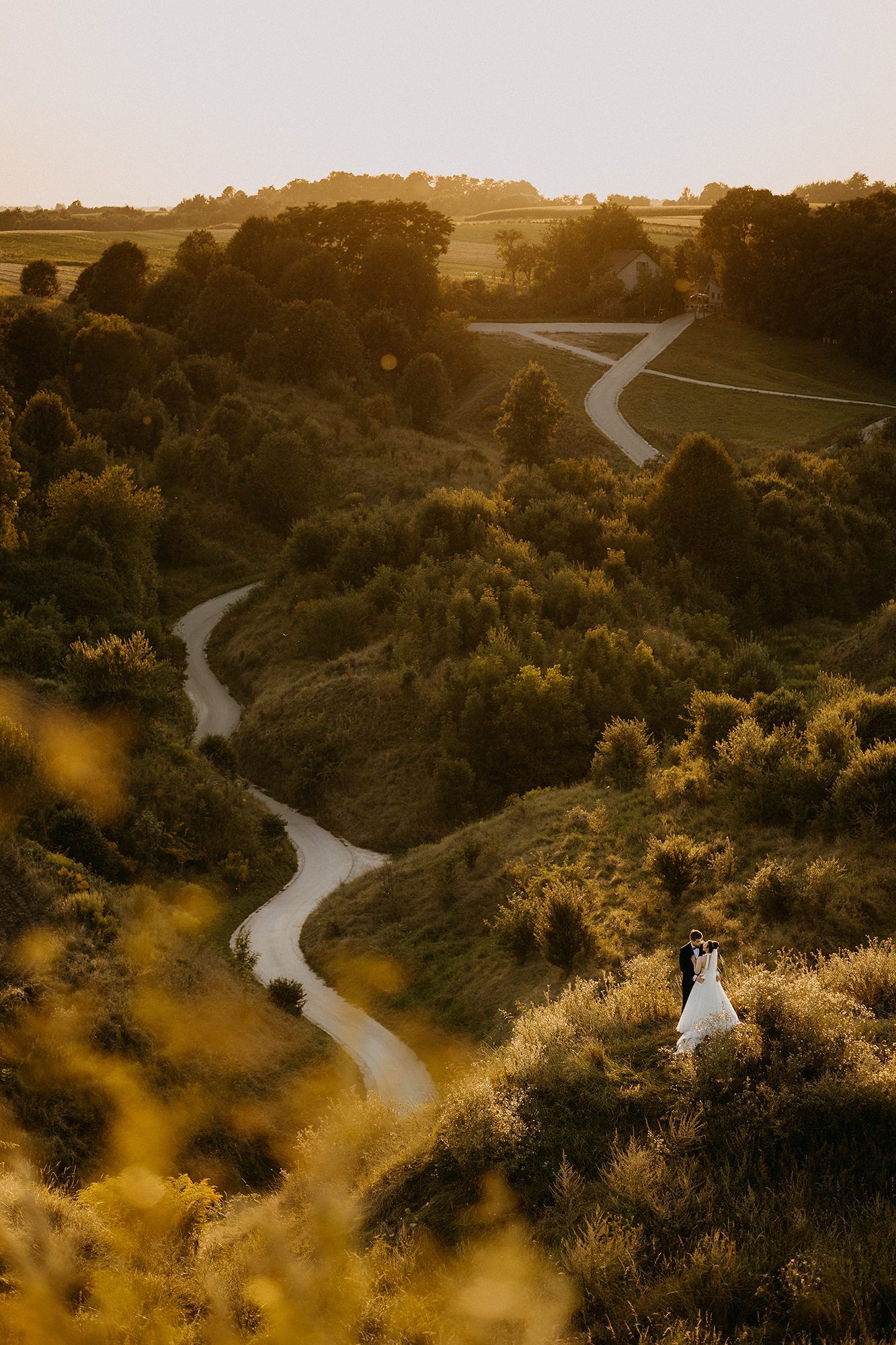 Aerial view of bride and groom on a sunlit hillside beside a winding country road.