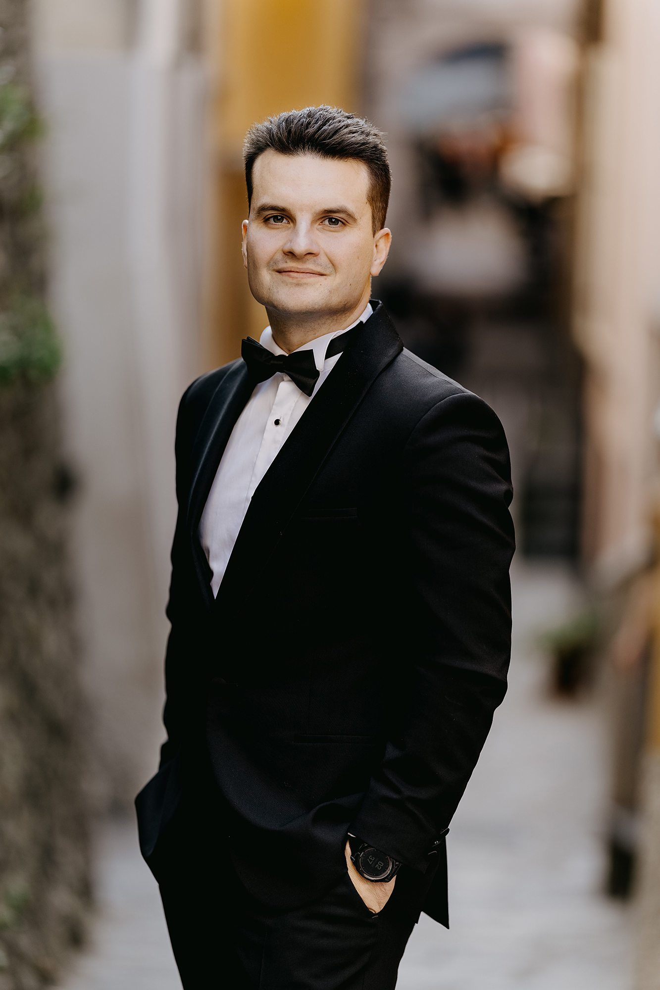 Dapper groom in black tuxedo and bow tie poses confidently in a softly blurred Italian alley.