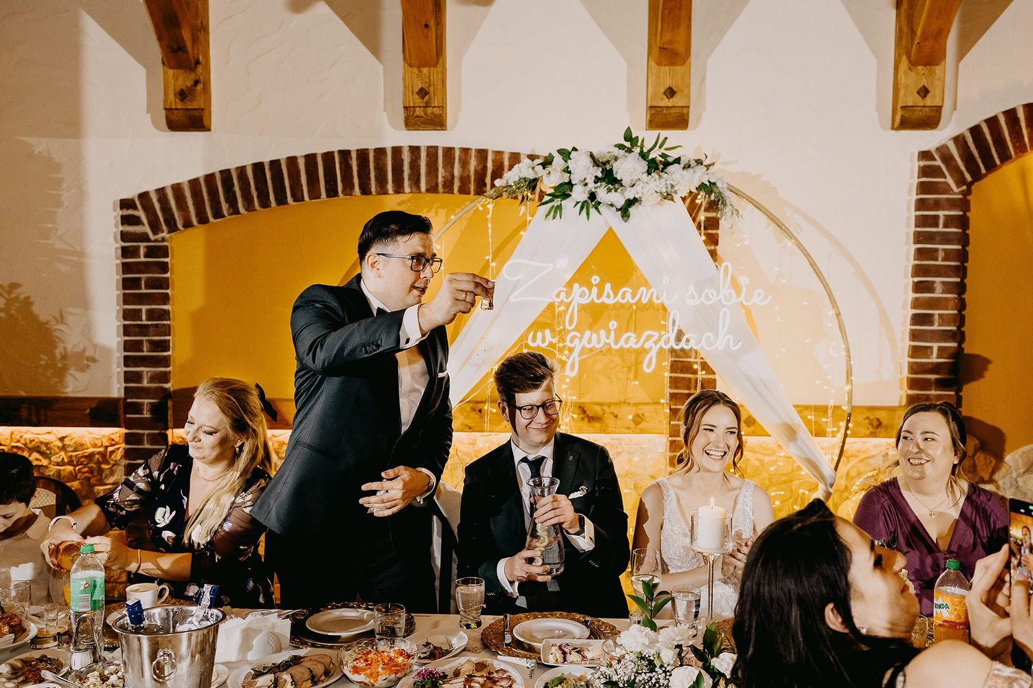 Wedding guest raises toast at reception dinner in rustic brick venue with neon sign backdrop.