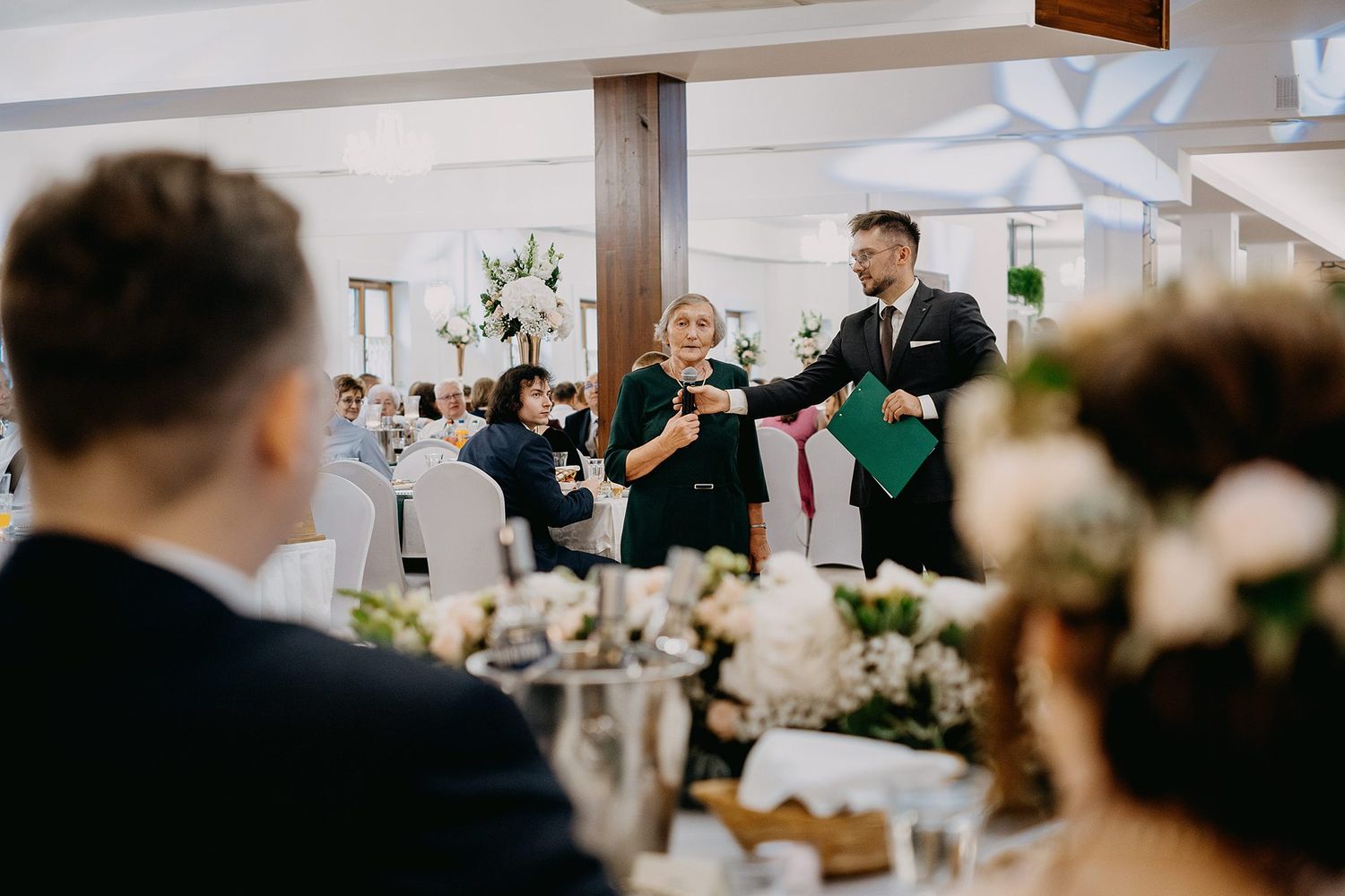 Wedding MC couple toasting guests at elegant reception venue with floral centerpieces.