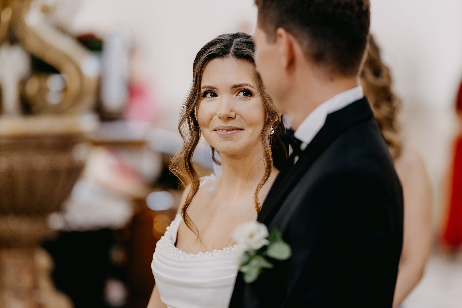 Bride gazing lovingly at groom during wedding ceremony, wearing an off-shoulder white gown with floral detail.