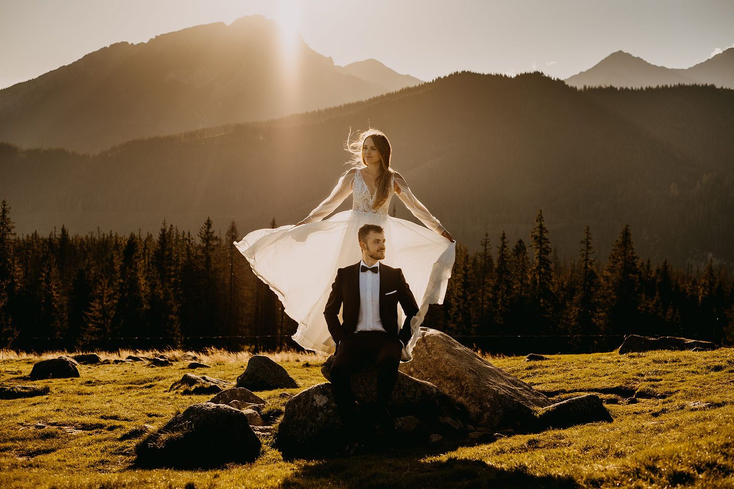 Bride stands above groom on mountain rocks with golden sunset and alpine peaks behind.