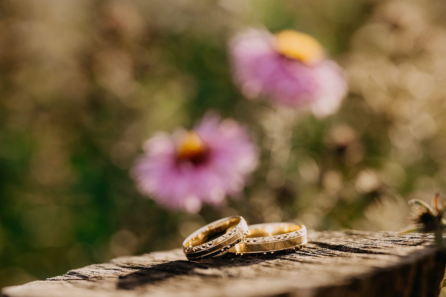 Two gold wedding rings resting on rustic wood with soft pink flowers in the blurred background.
