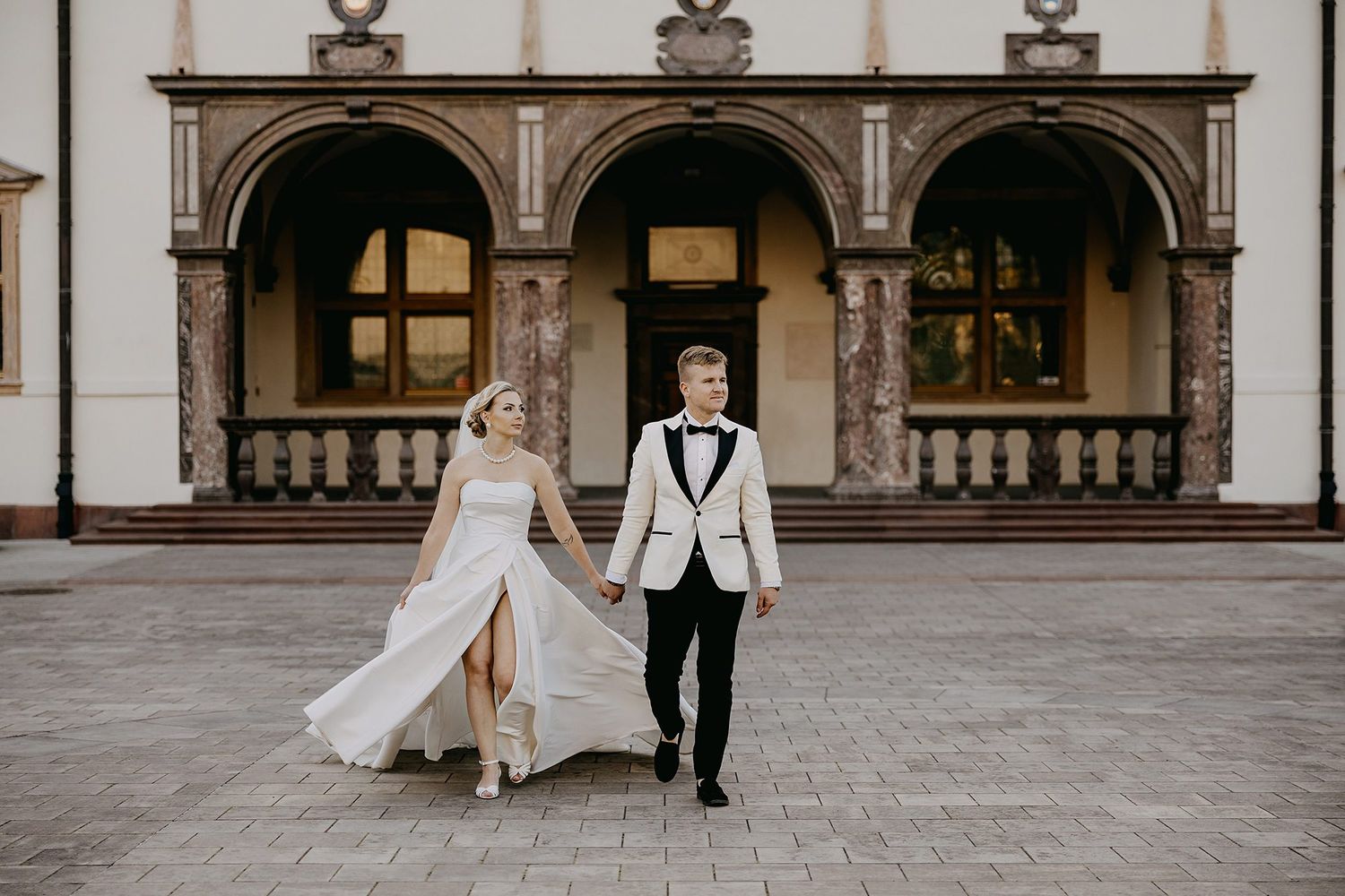 Elegant bride and groom holding hands walking in front of a grand building with arched columns.