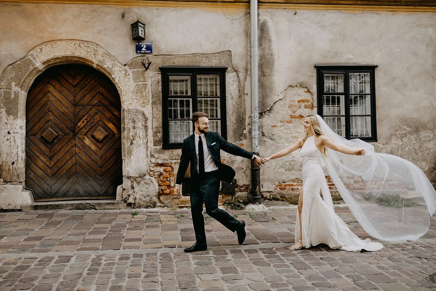 Bride and groom playfully running on cobblestone street with veil flowing in wind.