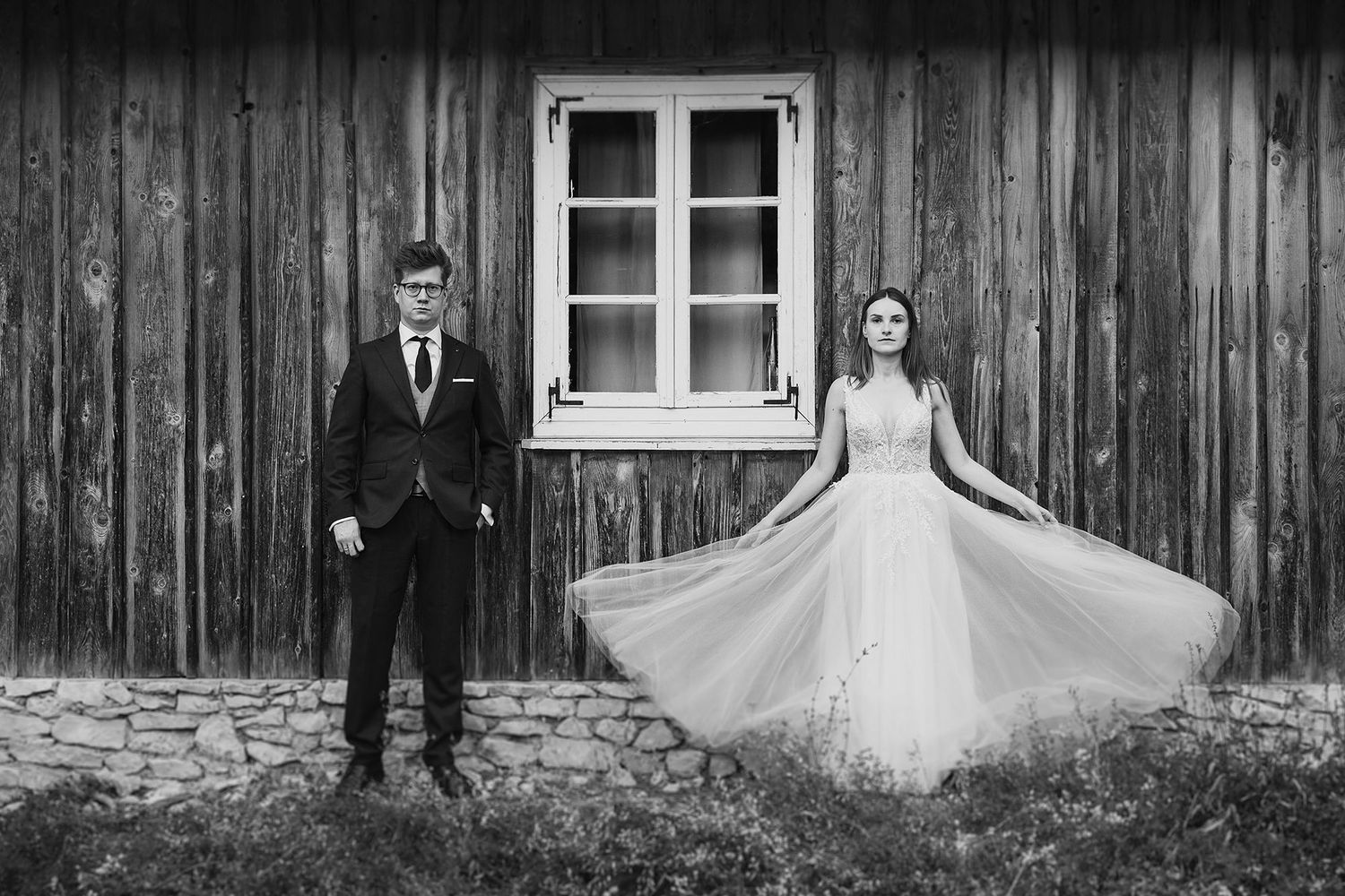 Black and white photo of bride and groom posing by a rustic wooden barn wall.