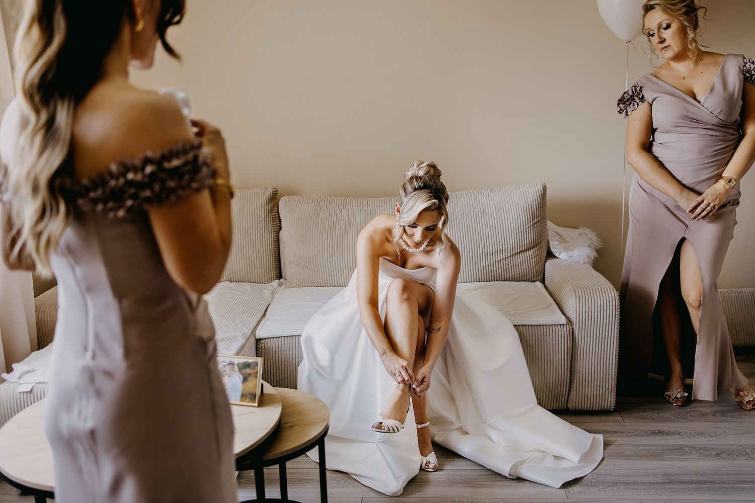 Bride adjusts her shoes while seated as bridesmaids assist during wedding preparations.