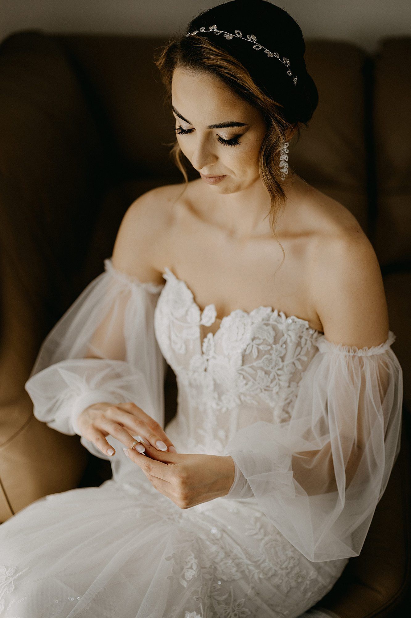 Elegant bride in off-shoulder lace wedding gown with headpiece admiring her ring while seated.