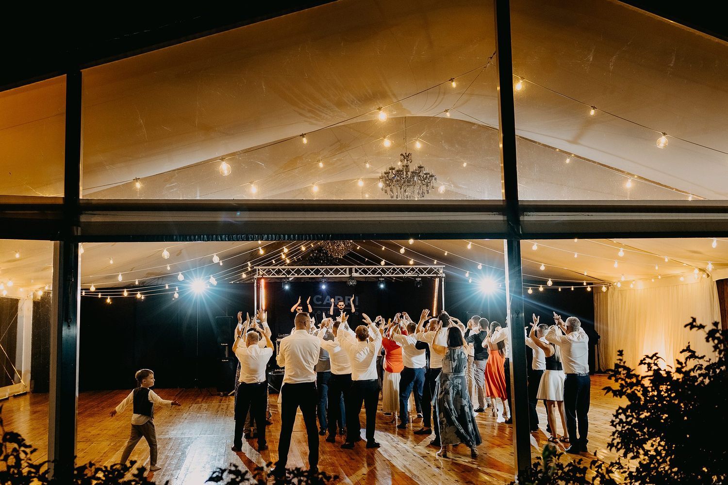 Guests dancing under string lights in a glass-walled wedding reception venue at night.