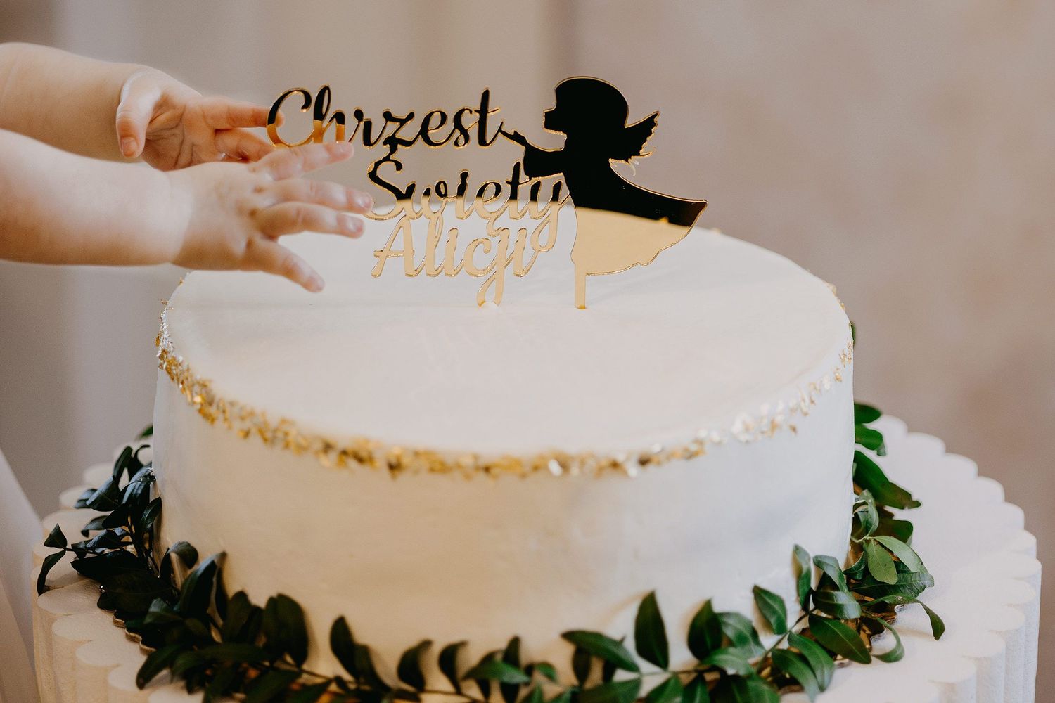 White baptism cake with gold angel topper reading Chrzest Swienty being decorated by hands.