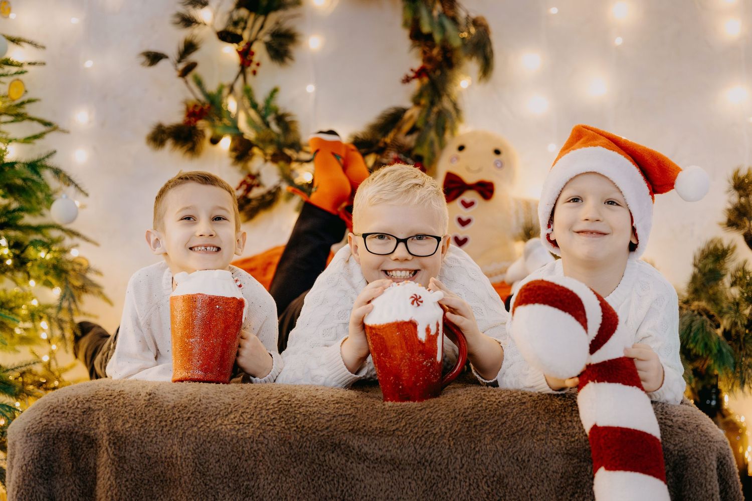 Three smiling children in Christmas pajamas and a Santa hat hold red cups of hot cocoa with whipped cream near festive garland and lights.
