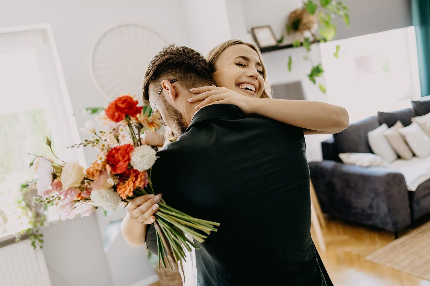 Happy bride hugging groom while holding a beautiful bouquet of orange and pink flowers indoors.