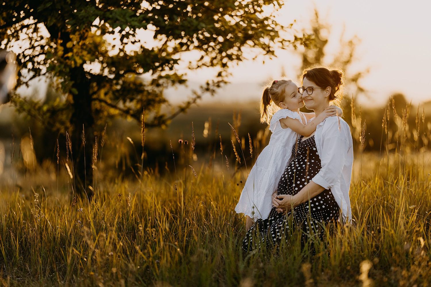 Pregnant mother in polka dot dress hugging young daughter in golden sunset meadow near trees.