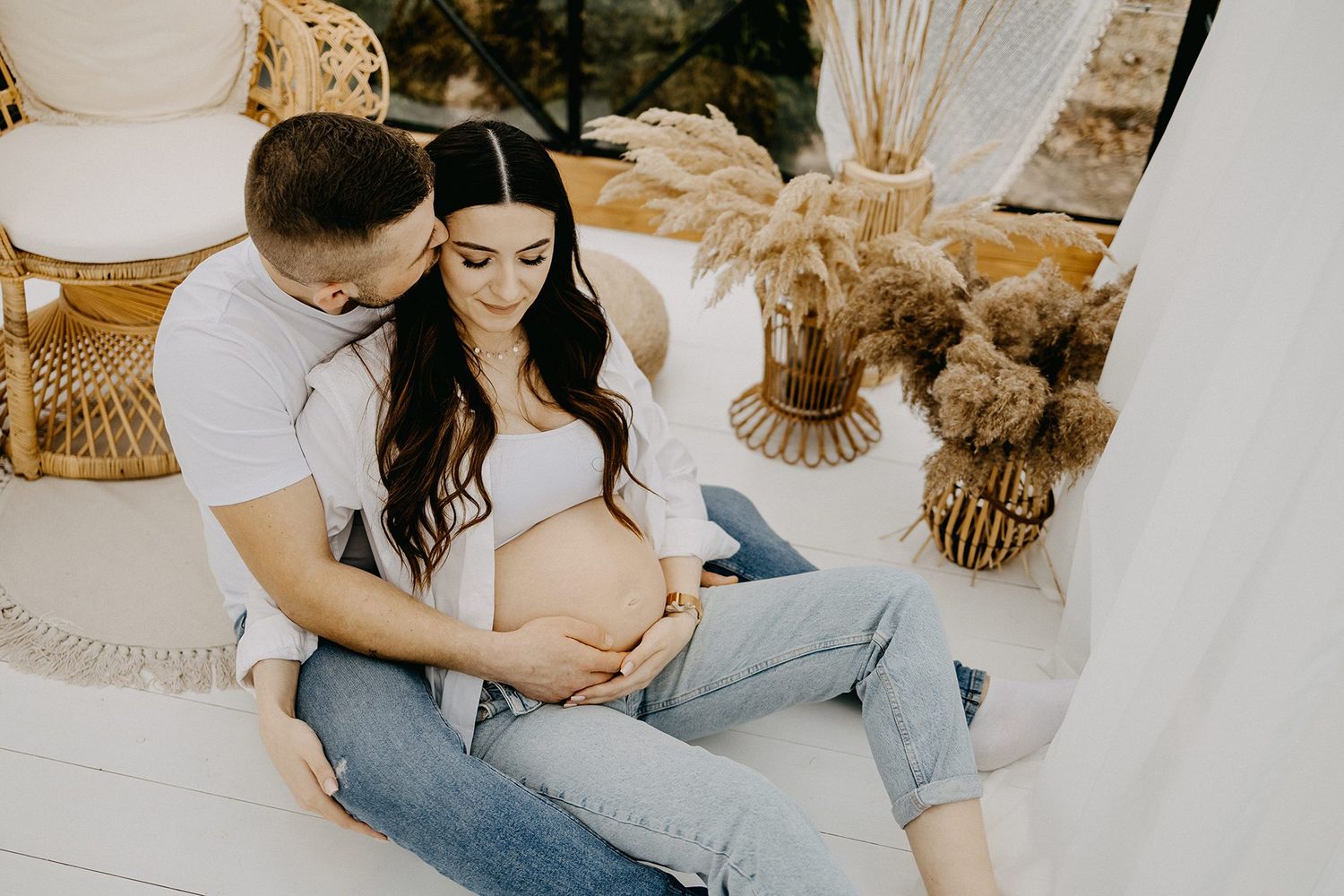 Couple sharing tender maternity moment, man embracing pregnant woman sitting on floor with boho decor.