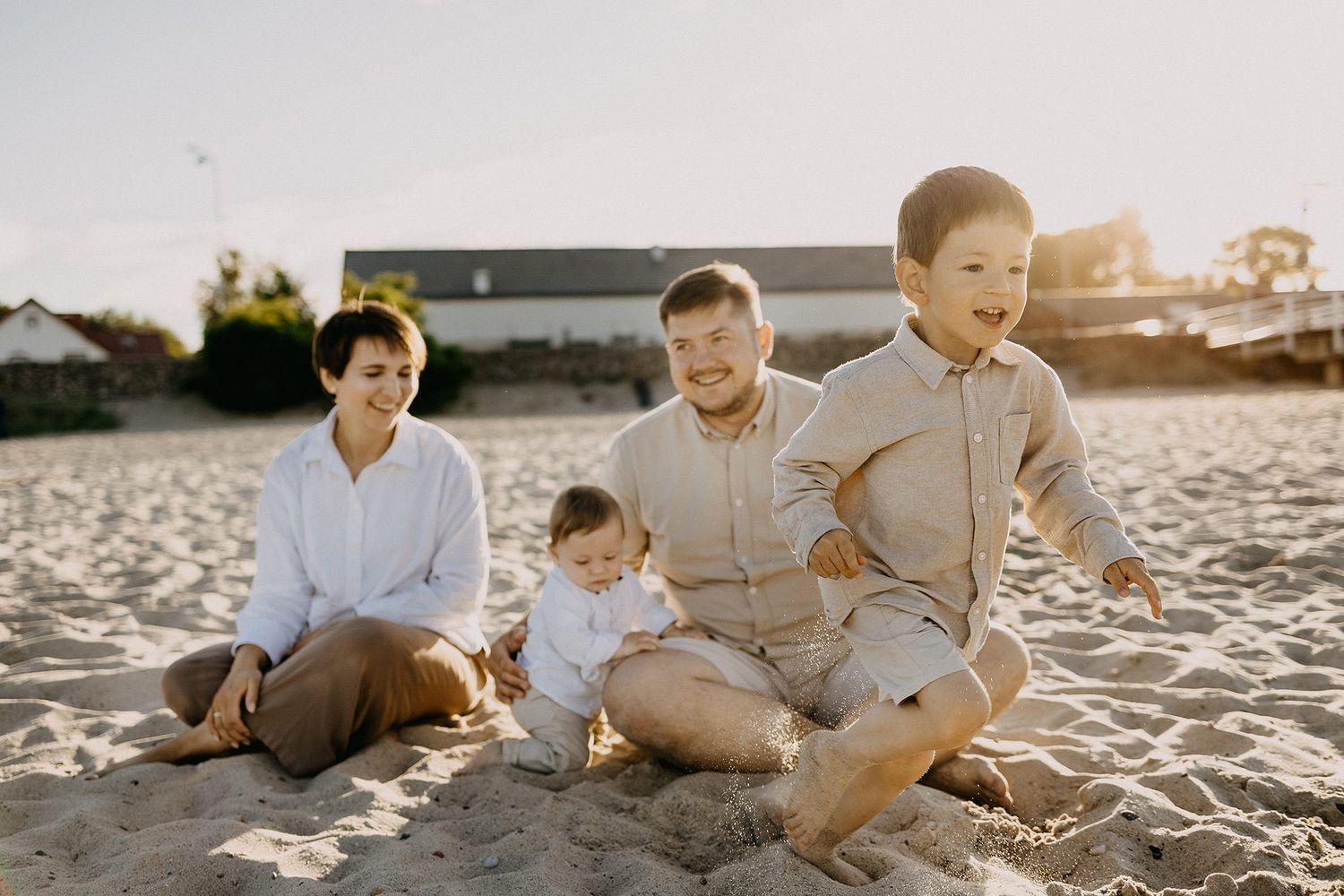 Family of four sitting on sandy beach at golden hour, parents smiling at baby while toddler runs nearby.