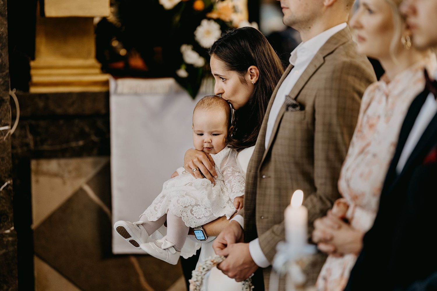 Parents holding baby in white christening gown during Catholic baptism ceremony inside ornate church.