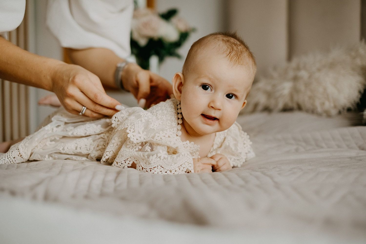Adorable baby in a white lace christening gown lies on a bed while an adult adjusts her dress.