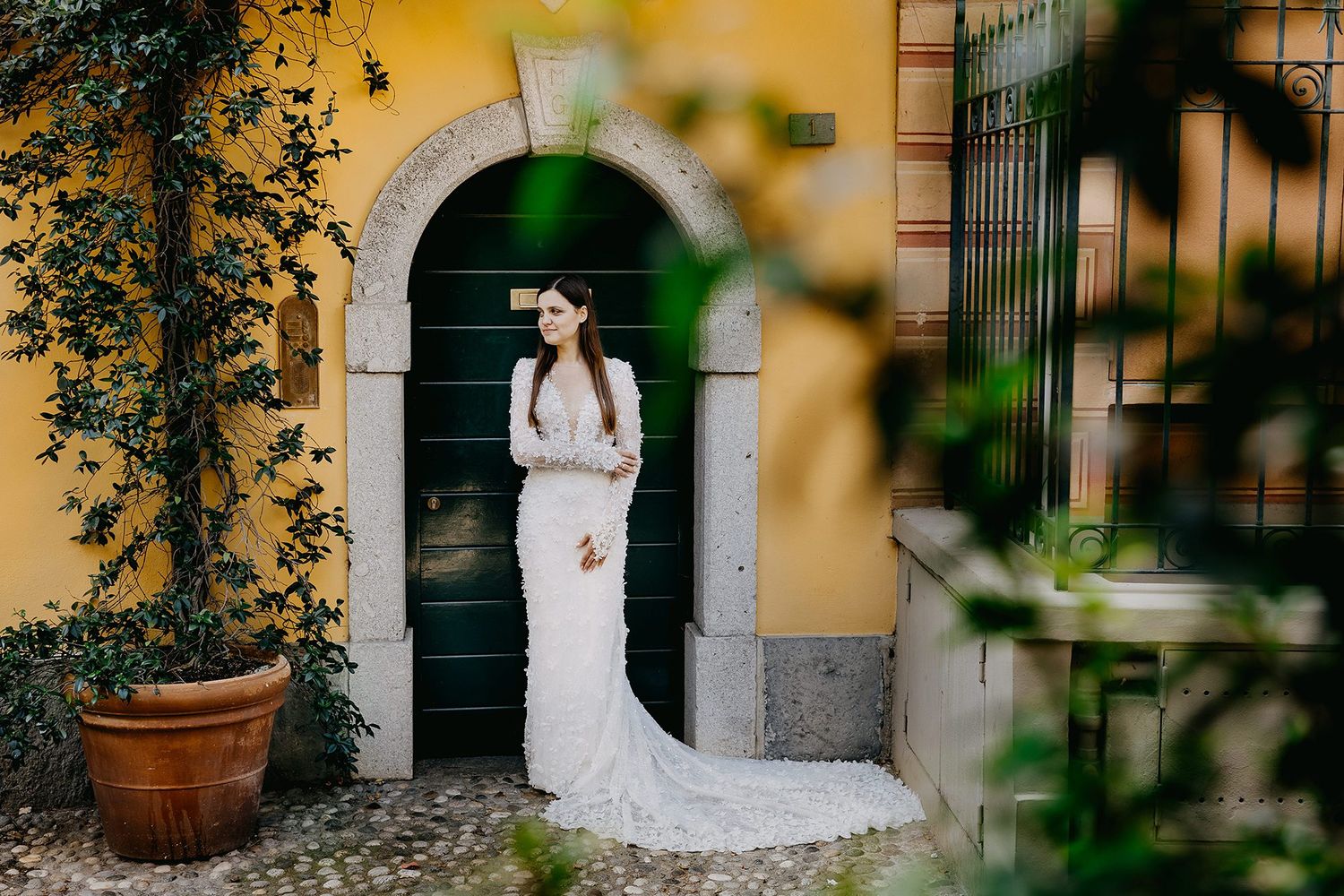 Elegant bride in long-sleeve lace wedding gown poses before a green door of a yellow Italian villa.
