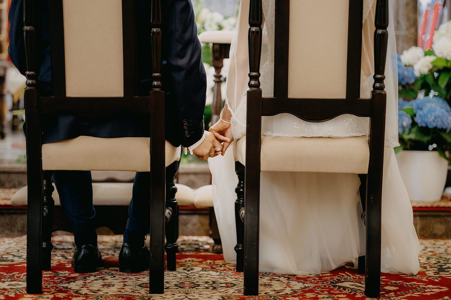 Bride and groom holding hands between ornate wooden chairs during a church wedding ceremony.