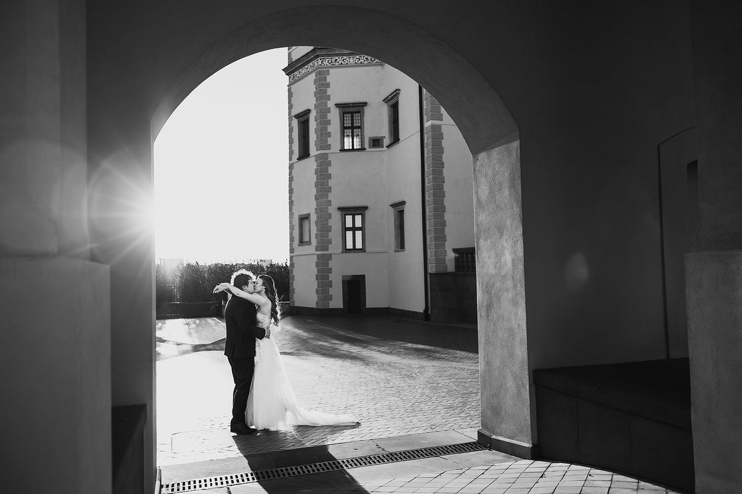 Black and white silhouette of bride and groom embracing under stone archway with sunburst backdrop.