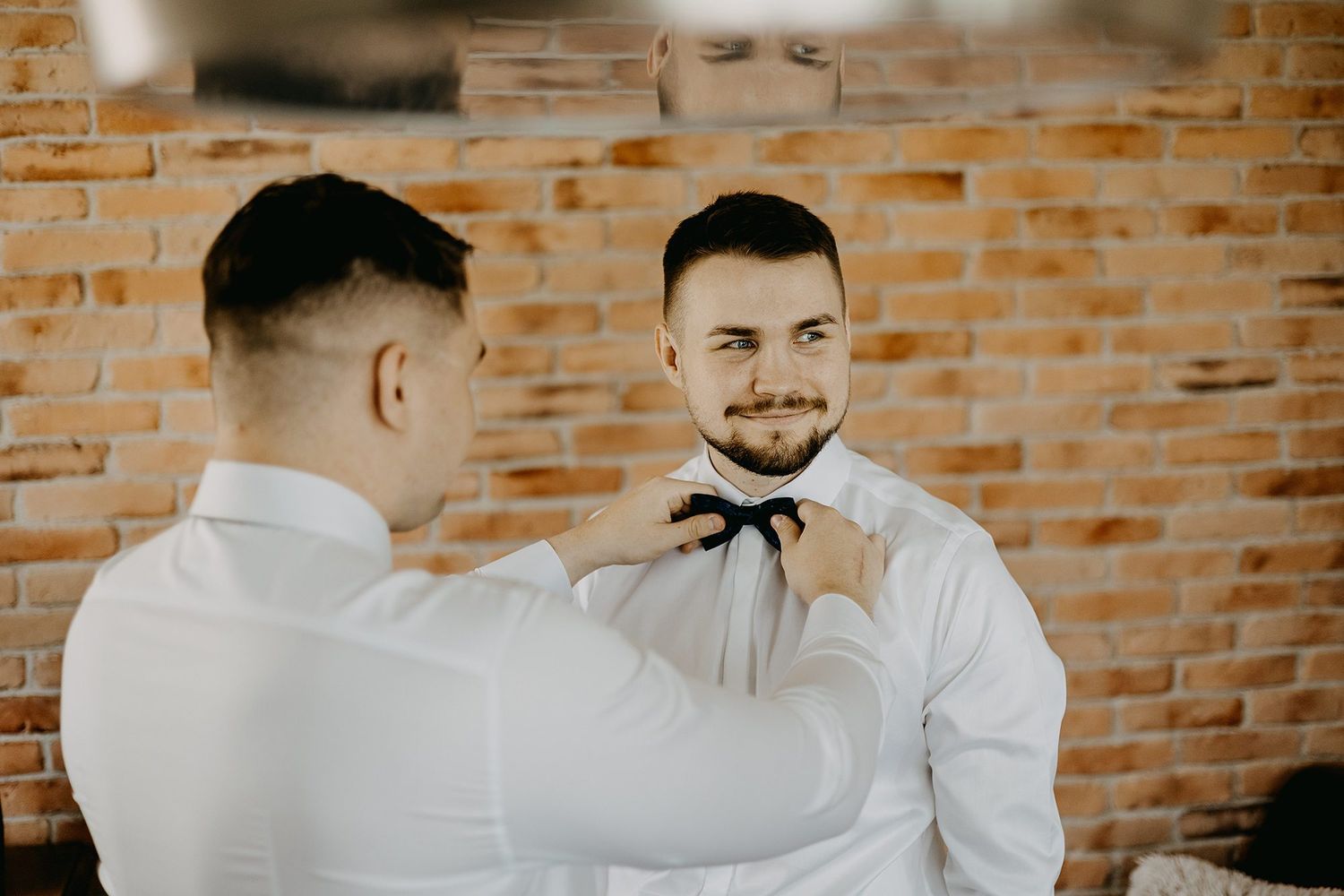 Best man helps smiling groom adjust black bow tie against exposed brick wall backdrop.