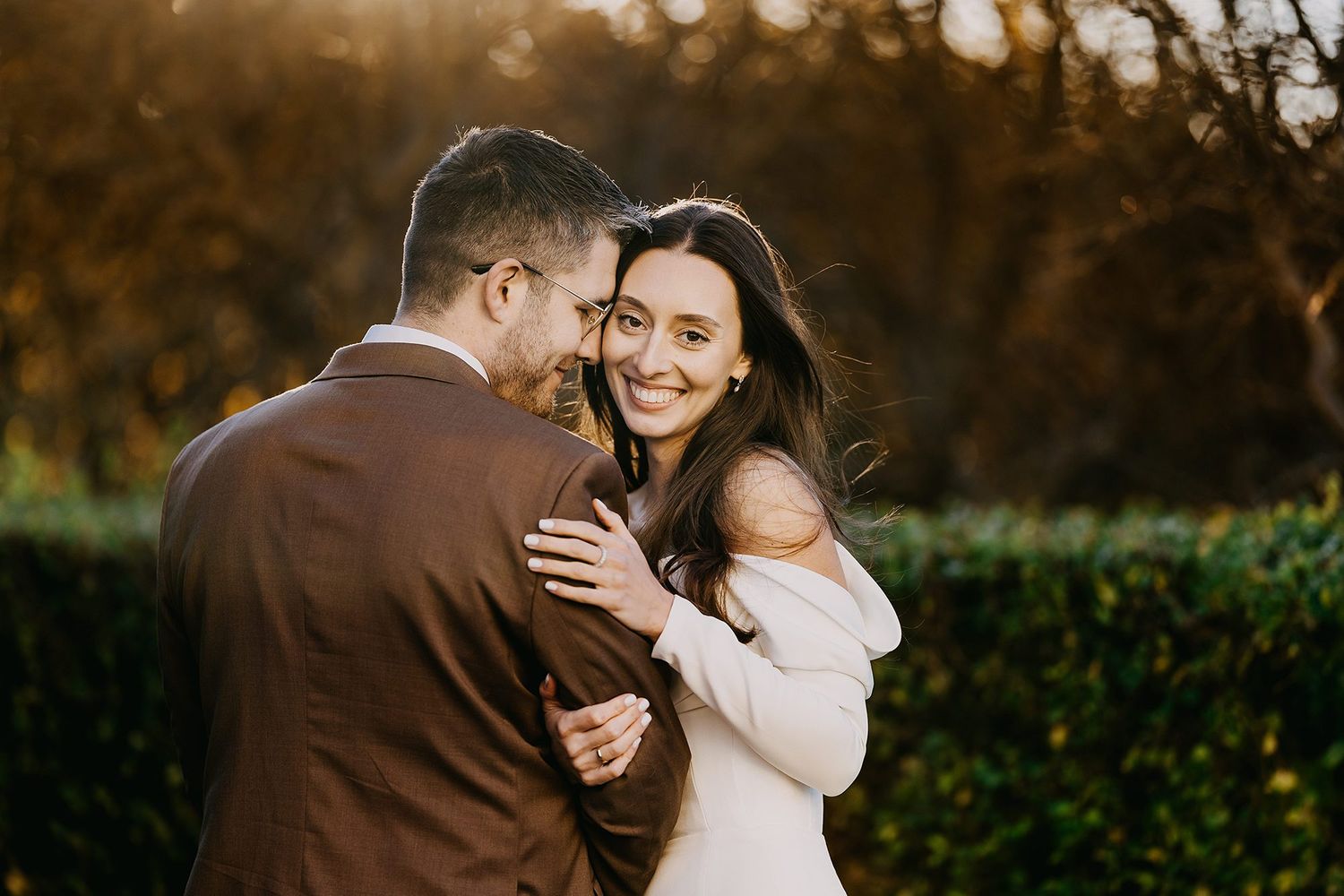 Smiling bride embracing groom in a sunlit garden at golden hour.