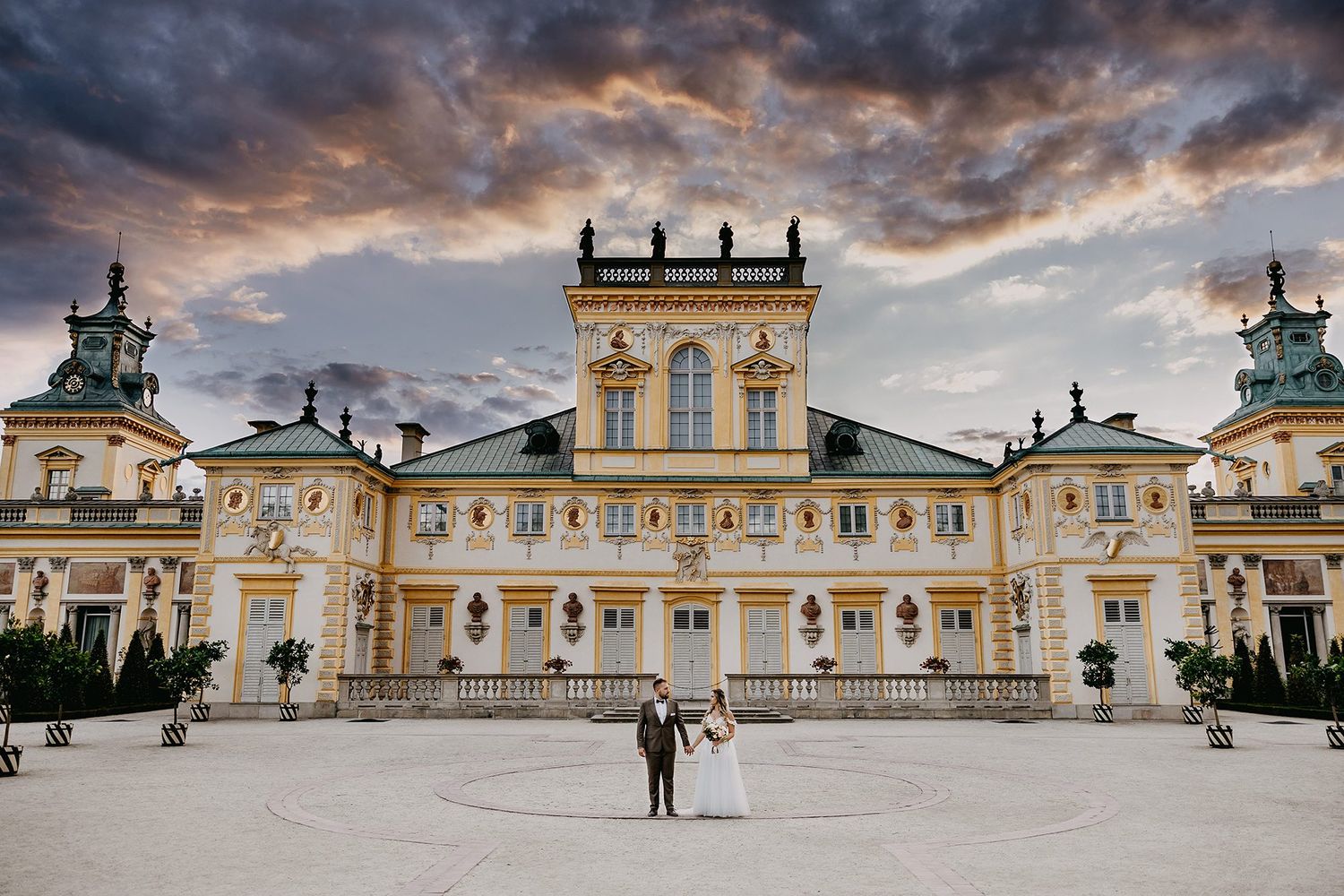 Bride and groom posing in front of an ornate Baroque palace under a dramatic cloudy sky.