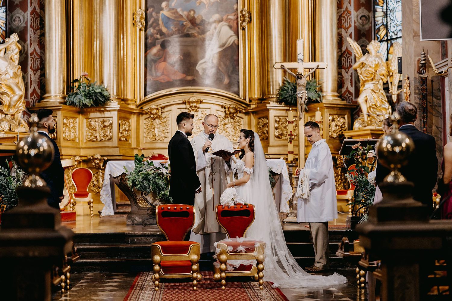 Bride and groom exchanging vows at elaborately decorated baroque church altar with priest officiating.