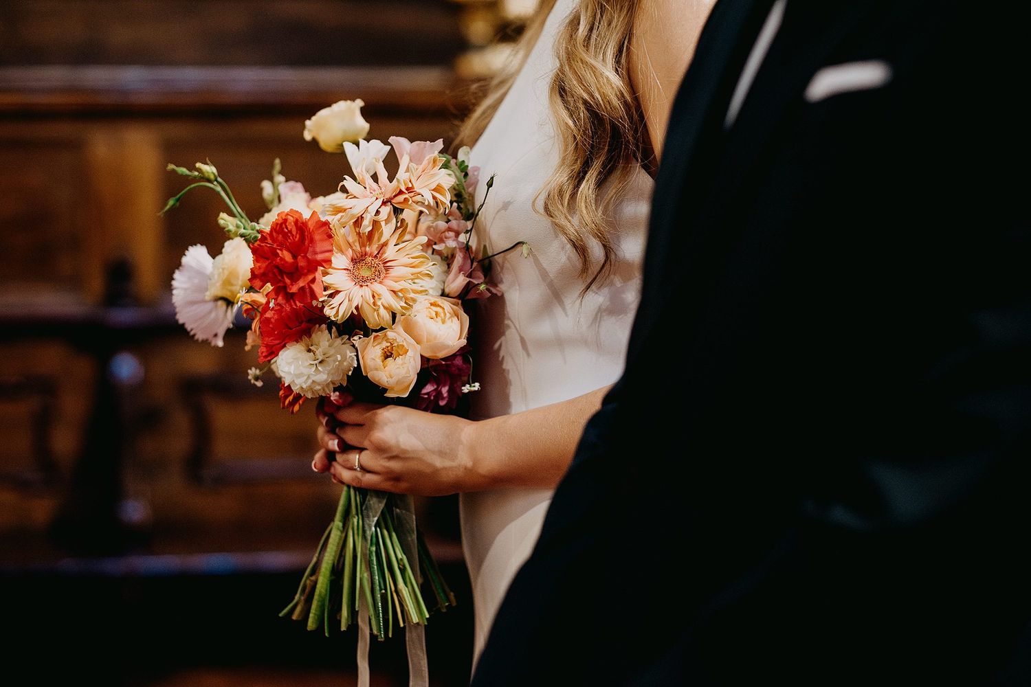 Close-up of a bride holding a vibrant bouquet of dahlias and ranunculus in warm orange and pink tones.