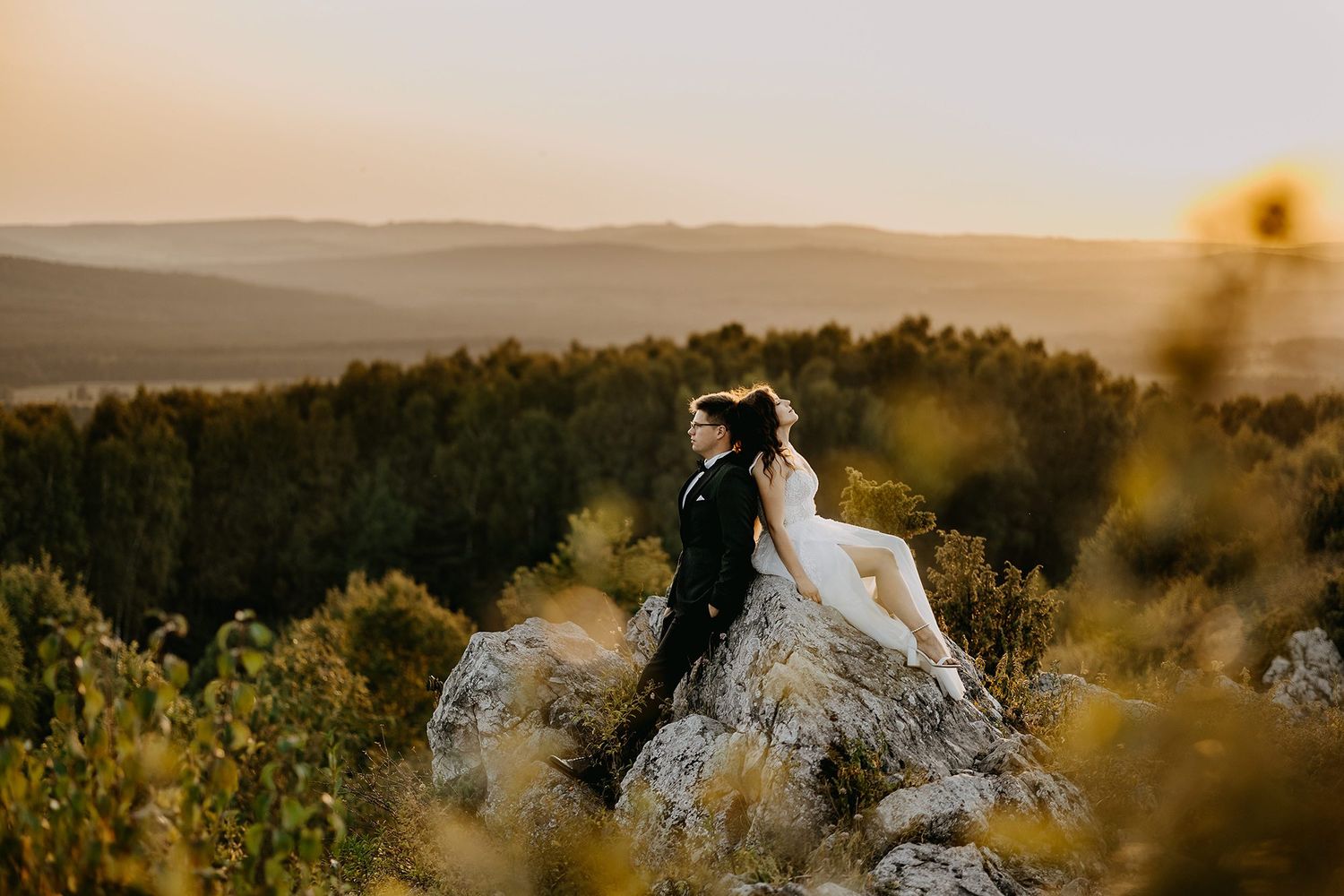 Wedding couple sits on a rocky hilltop embracing at sunset overlooking a vast scenic landscape.
