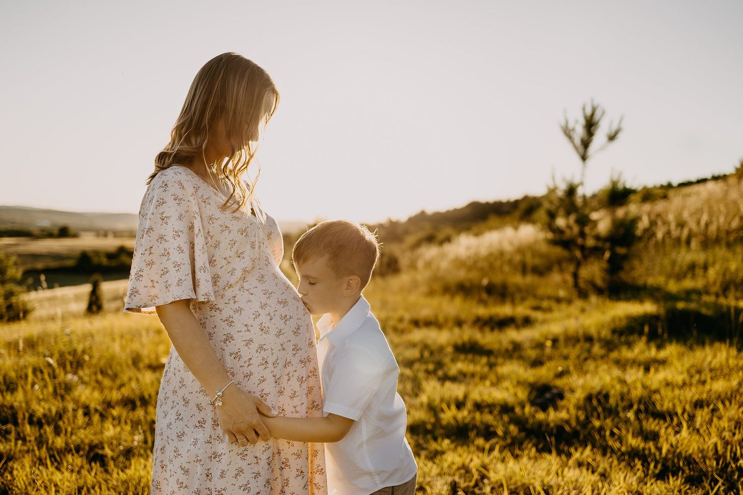 Young boy hugs pregnant mothers belly in a sunlit golden meadow during maternity photoshoot.