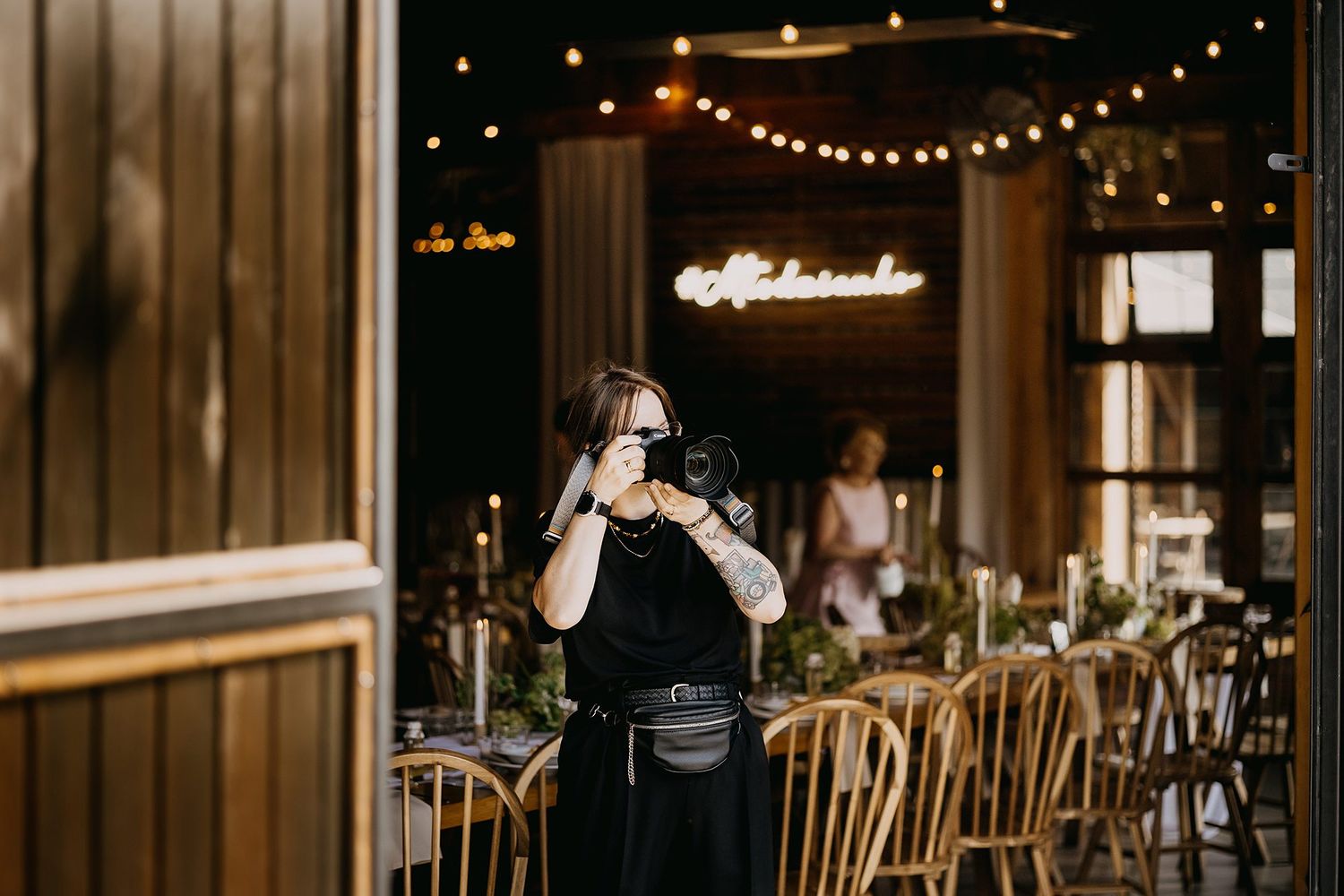 Female photographer taking photos with a DSLR camera inside a warmly lit rustic wedding venue.