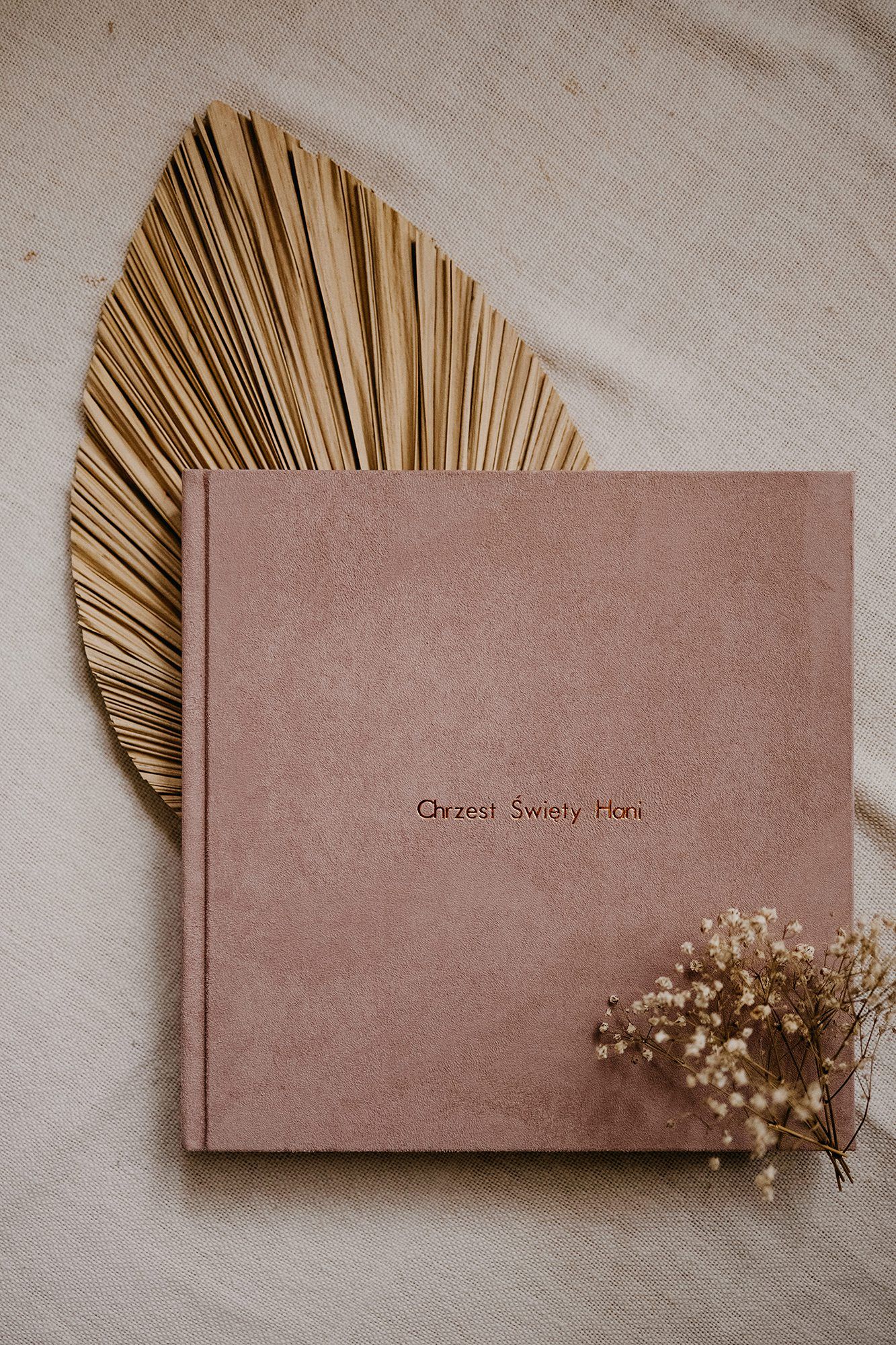 Dusty rose velvet photo album with gold text on white linen beside dried palm leaf and flowers.