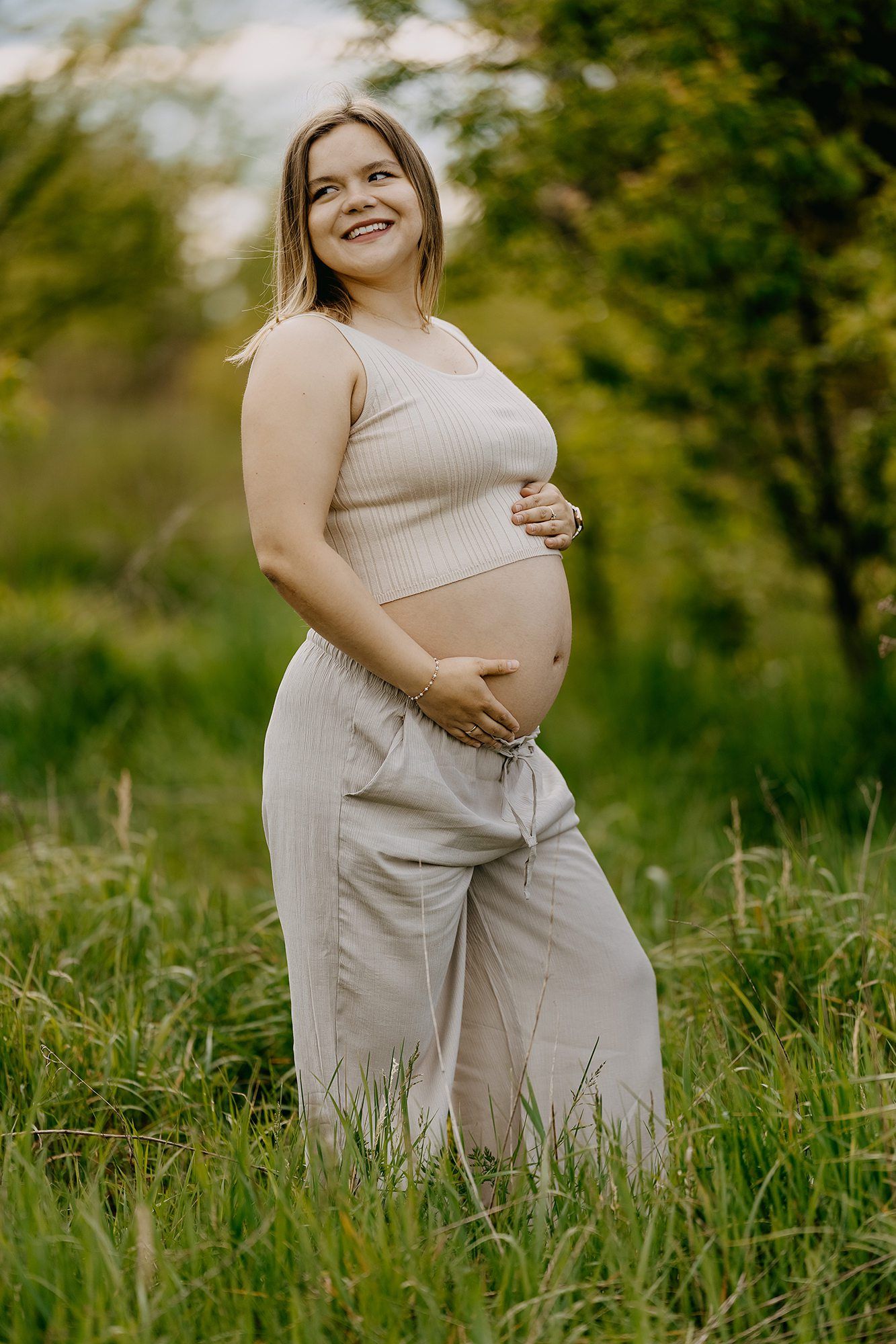 Smiling pregnant woman in beige crop top and pants cradling her baby bump outdoors in green field.