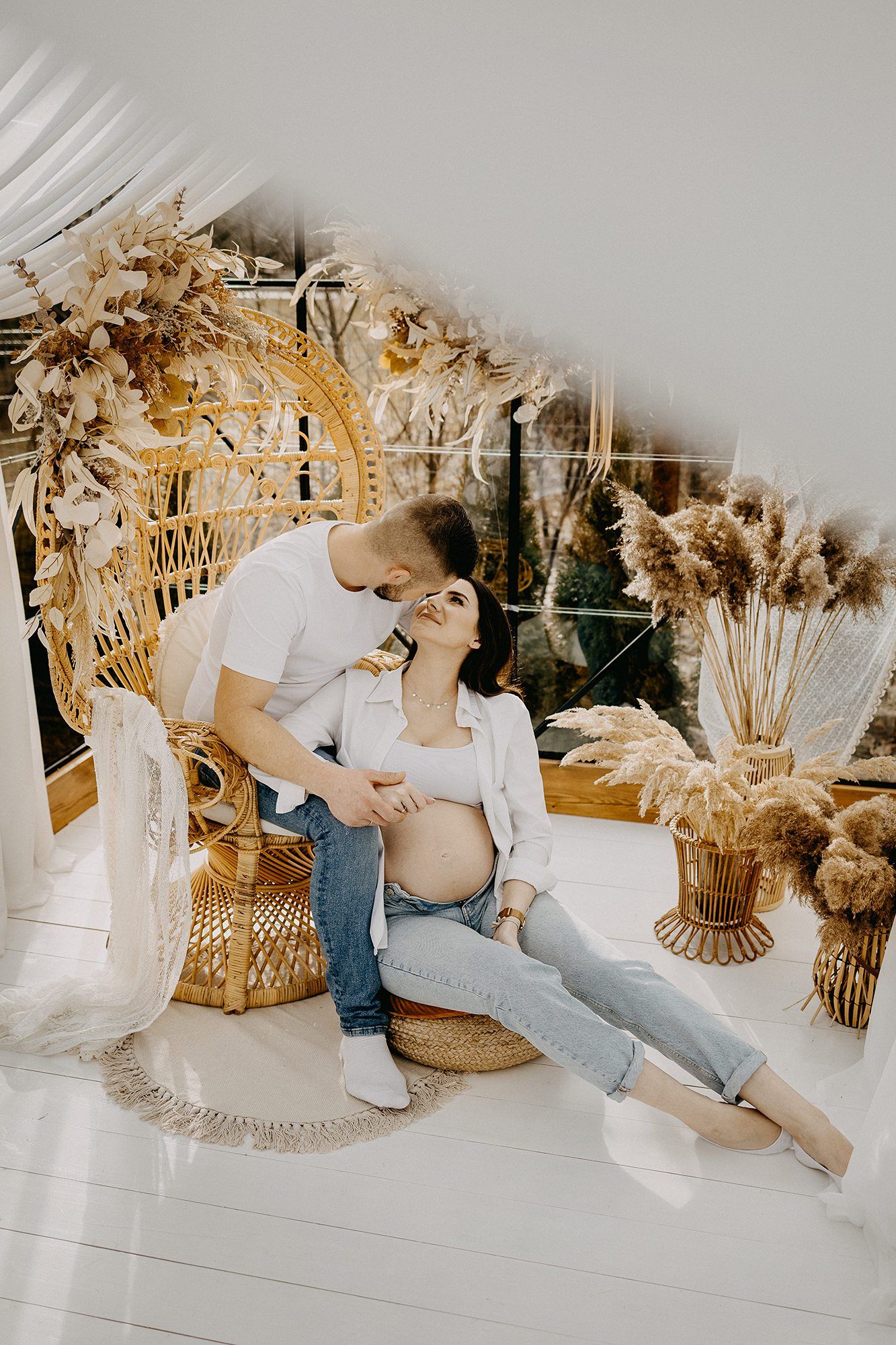 Loving couple in boho-styled studio with man kissing pregnant womans head near rattan peacock chair.