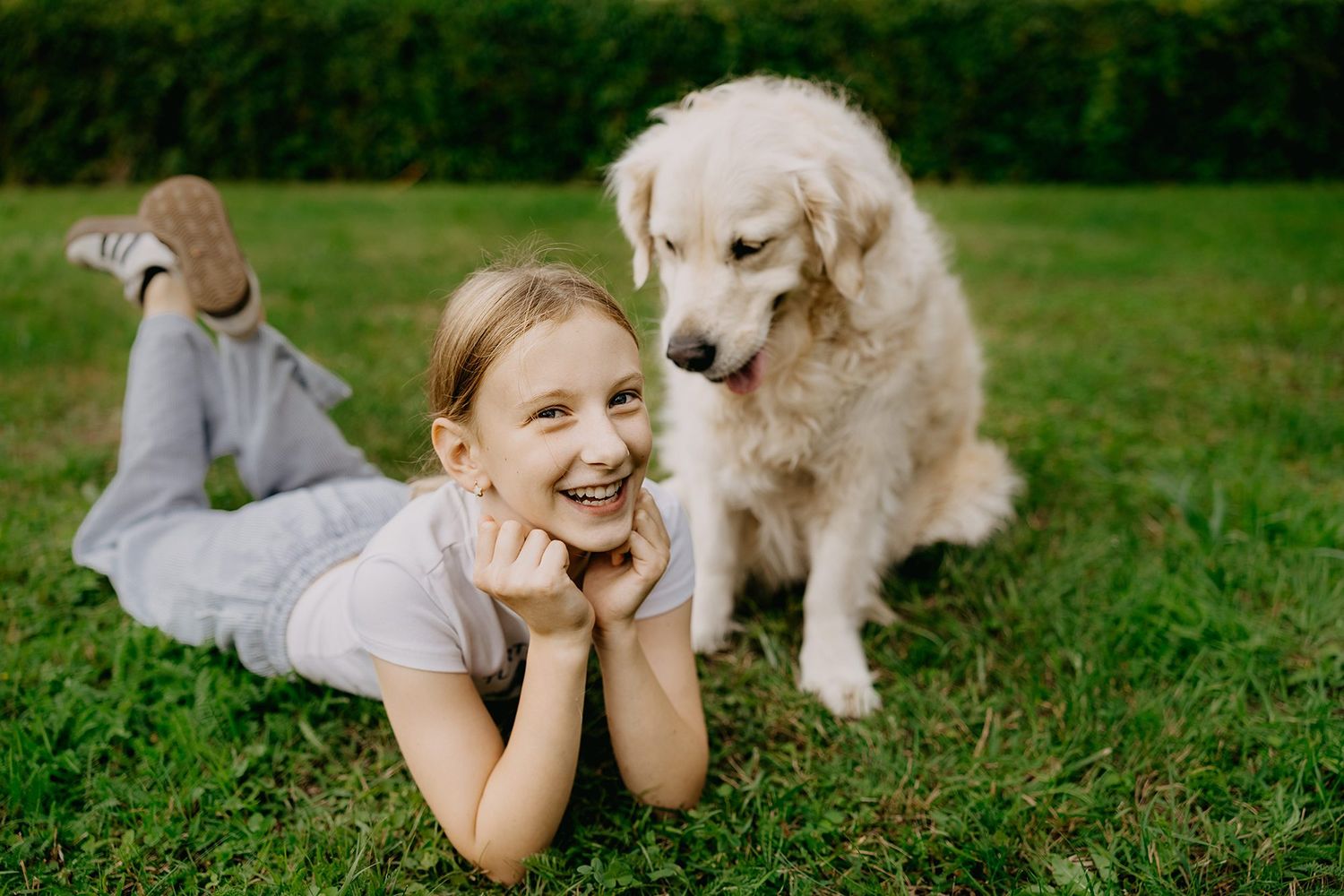 Smiling girl lying on green grass with a fluffy white Golden Retriever dog beside her outdoors.