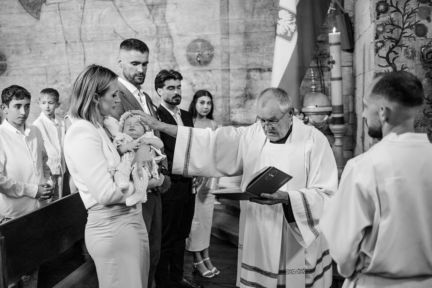 Black and white photo of a Catholic baptism ceremony with priest blessing an infant held by the mother.